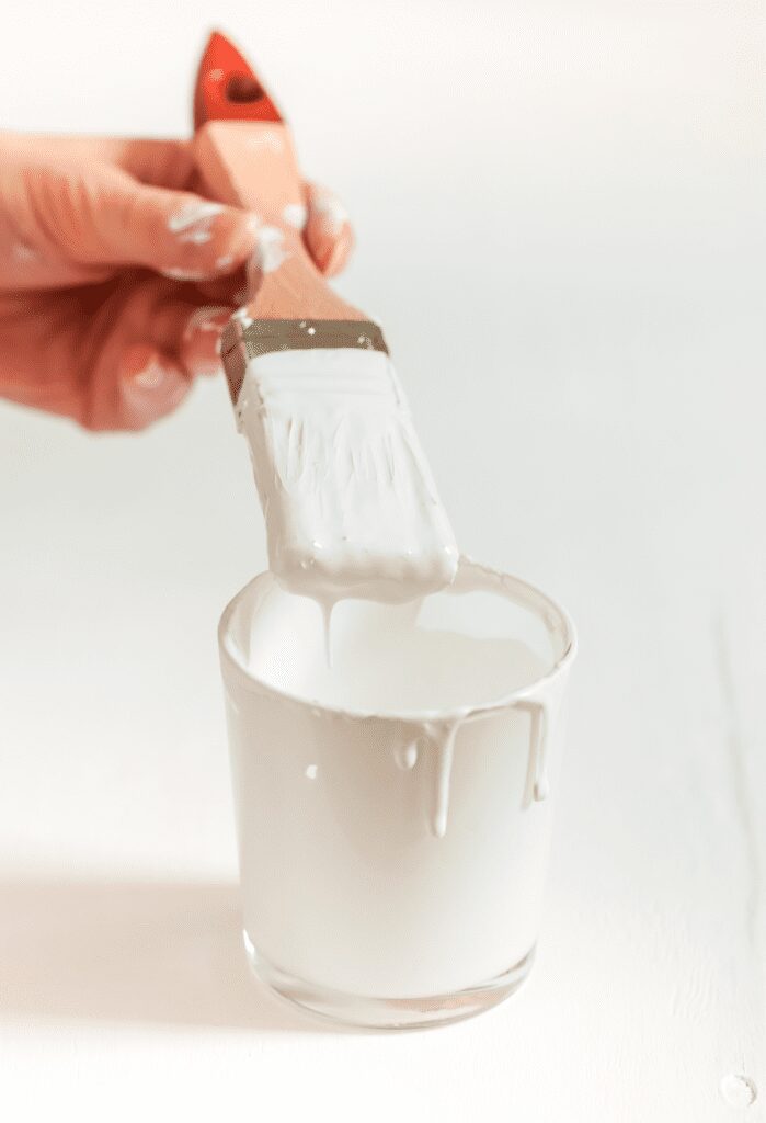 A close-up of an artist’s hands dipping a wide brush into a jar of acrylic gesso, preparing to prime a canvas.